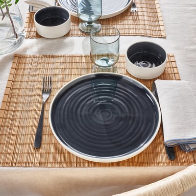Table avec des plats minimalistes OMBONAD. Comprend une assiette en céramique, de petits bols, du verre, une fourchette et une serviette sur un tapis tissé.