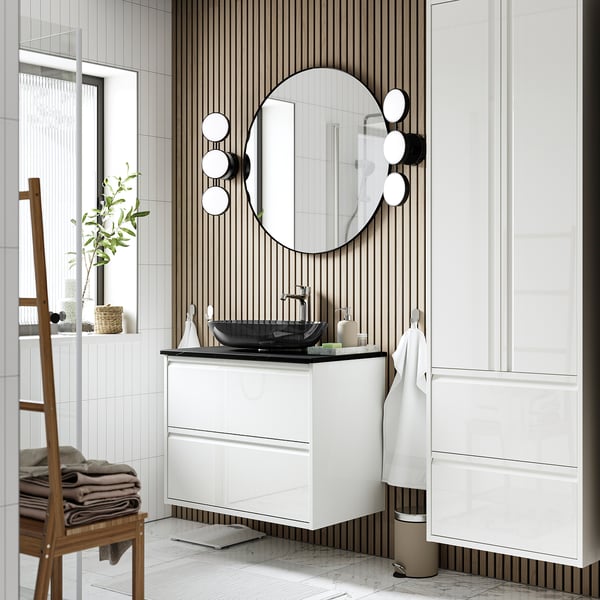 Salle de bain moderne avec lavabo en verre gris foncé OXMYREN. Grand miroir rond, armoires blanches, chaise en bois et mur rayé. Design élégant et flottant.
