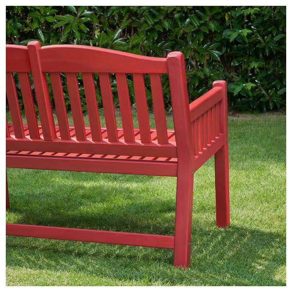 Un banc PÄRONHOLMEN rouge en bois d’acacia. Le banc a un dossier incurvé et des lattes droites, conçu pour une utilisation en extérieur, posé sur une pelouse herbeuse.