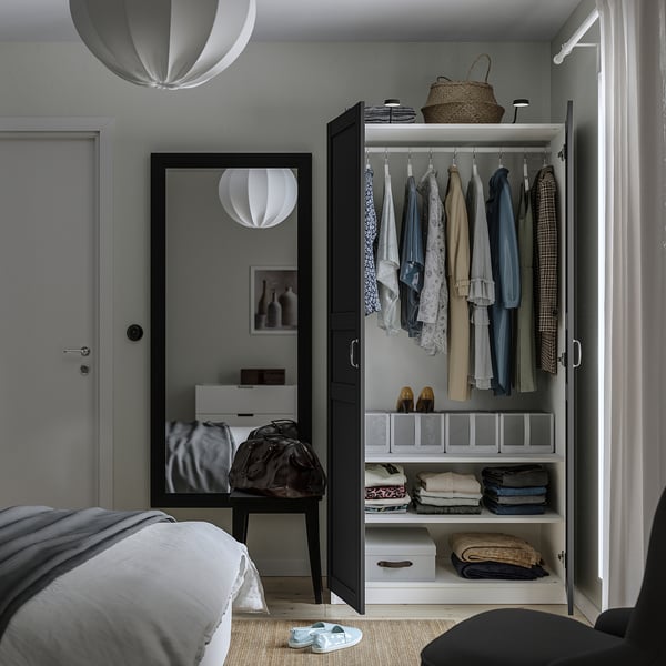 Chambre avec armoire ouverte exposant des vêtements, des vêtements pliés et des boîtes de rangement. Le miroir sur la porte reflète le décor de la pièce.