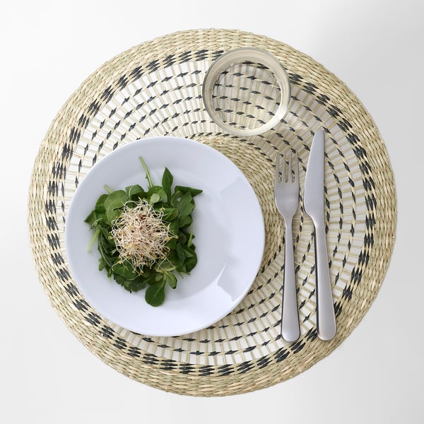 Un napperon rond tissé avec un motif gris et blanc contient une assiette blanche de salade et de couverts.