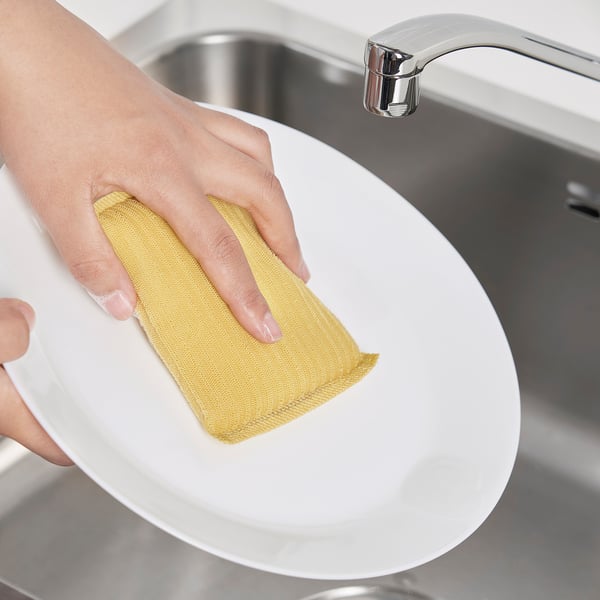 Personne nettoyant une assiette blanche avec un tampon à récurer jaune sous un robinet chromé.