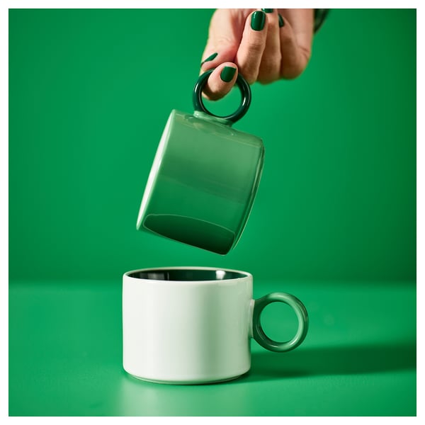 Une main tient une tasse PIGGÅL verte au-dessus d’une blanche. La tasse verte a une poignée ronde unique, un design simple et un glaçage coloré.