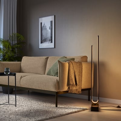 Salon moderne avec lampadaire PILSKOTT, canapé beige et plante. La lampe a un design noir élégant et haut avec un éclairage chaud.