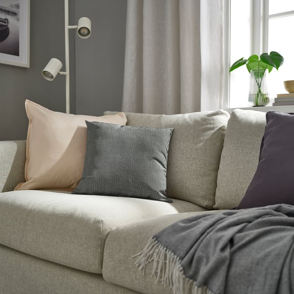Salon moderne avec coussin PROFETBLOMMA sur canapé gris près de la fenêtre avec rideaux gris clair. Décor écologique.