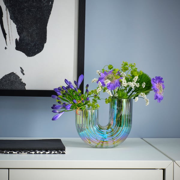 Un vase RÄFFELBJÖRK aux allures d’arc-en-ciel avec des fleurs violettes, sur une table blanche à côté d’un livre et d’une photo.