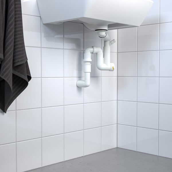 Évier blanc avec siphon d’eau RÄNNILEN visible et tuyauterie flexible contre le mur carrelé.