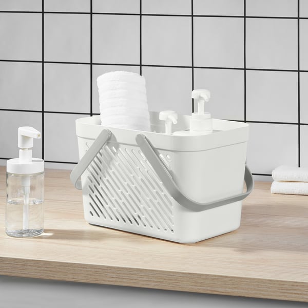 Panier de douche en plastique blanc avec poignées pliables, empilé avec des serviettes blanches roulées, posé sur un comptoir en bois clair à côté d’un distributeur de savon transparent.