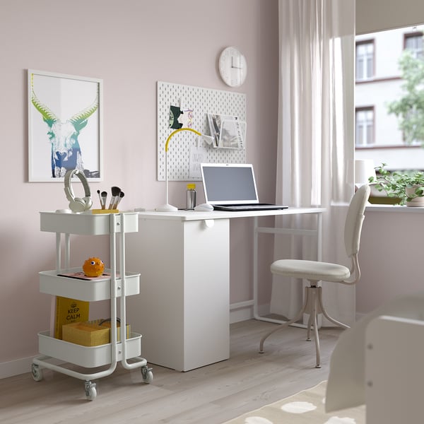 Espace de travail compact aux parois roses avec chariot RÅSKOG, ordinateur portable, écouteurs, tasse, conteneurs et panneau perforé. Éclairé par une grande fenêtre et une chaise blanche.