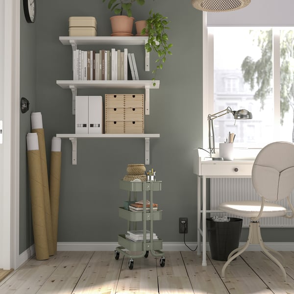 Chariot RÅSKOG vert et étagères blanches dans le bureau à domicile. Permet de ranger des objets, de se glisser sous les bureaux. La chambre a du parquet clair et un bureau bien éclairé.