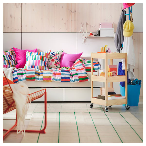 Chambre lumineuse et colorée avec canapé rayé, oreillers et tapis, chariot de rangement en bois, chaise rouge et objets suspendus.