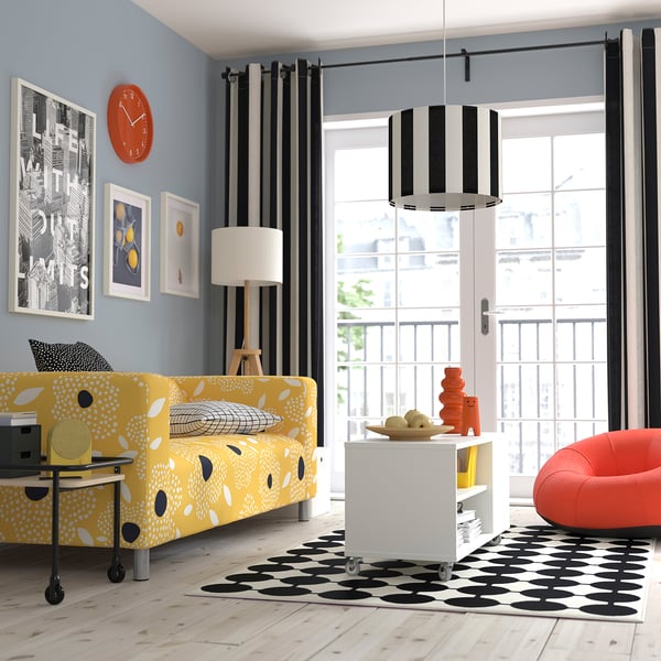 Canapé RINGSTA jaune, tapis noir/blanc, horloge orange, rideaux rayés. Grande fenêtre avec balcon, lampe moderne au-dessus d’une petite table avec des vases orange.