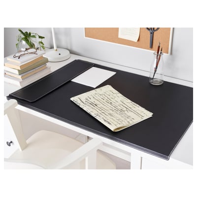 Le sous-main noir protège le bureau blanc, reste en place avec un bord lesté. Entouré de livres, d’une lampe, d’un vase à stylo et d’un tableau en liège.