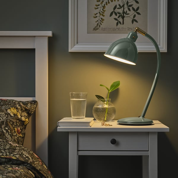 Chambre confortable avec lampe de bureau RÖDFLIK verte sur une petite table à côté du lit, avec abat-jour réglable, verre d’eau et petit vase à plantes.