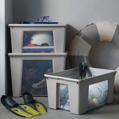 Trois boîtes de rangement SAMLA grises, fenêtres transparentes, divers équipements de sport à l’intérieur, empilés, plus grands au sol, plus petits sur le dessus.