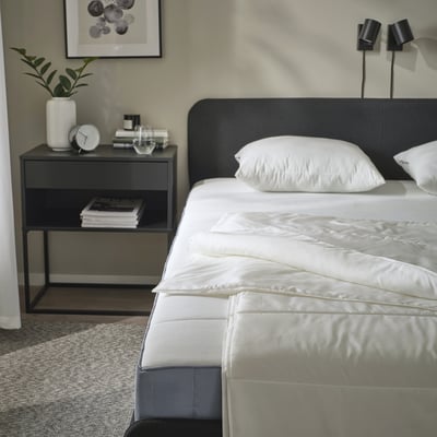 Scène de chambre à coucher avec couette SANDGRÄSMAL sur le lit, table de chevet grise contenant des livres, une horloge et une plante ; tête de lit noire, appliques murales.