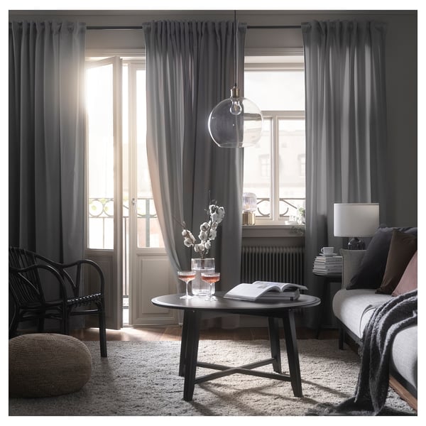 Salon moderne avec rideaux gris foncé, chaise en osier, table en bois, verres à vin et tapis. Grande fenêtre avec lumière douce.