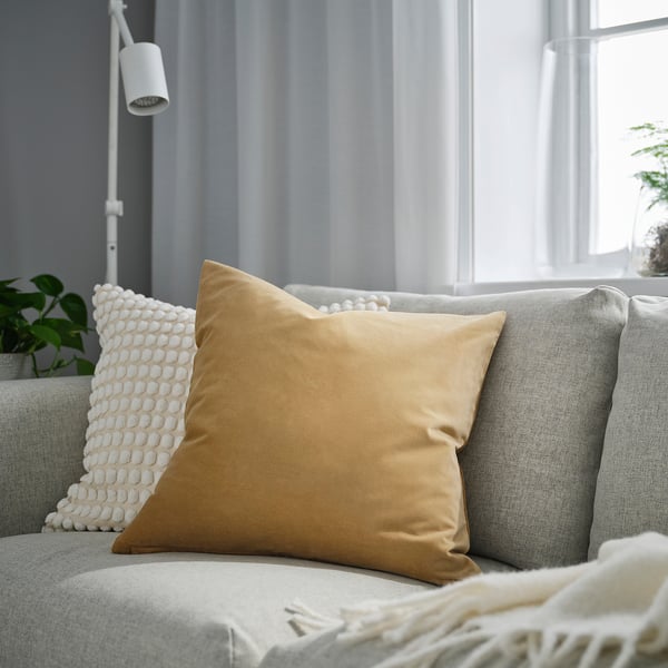 Salon avec un grand coussin SANELA jaune moutarde sur un canapé gris clair, accompagné d’un coussin à texture blanche, près d’une fenêtre à rideau blanc.
