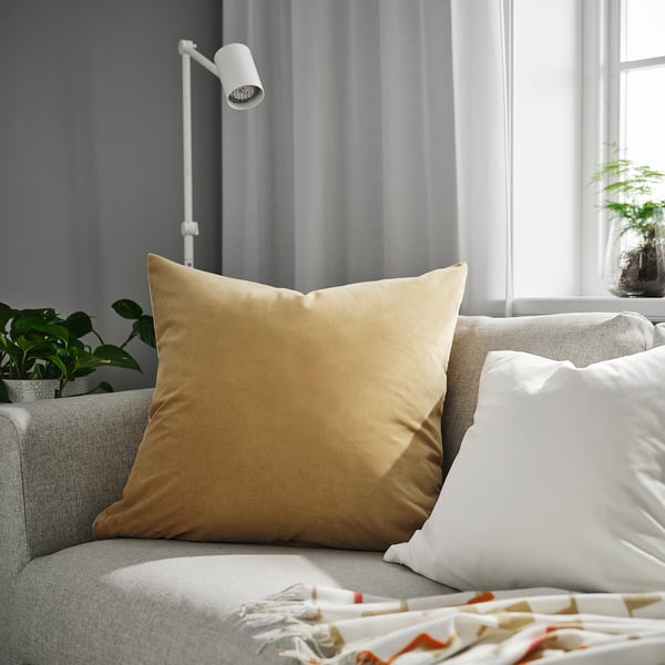Grand coussin SANELA doré sur canapé beige à côté d’un oreiller blanc, d’une lampe moderne, d’une plante verte et d’un lit avec une couverture colorée.