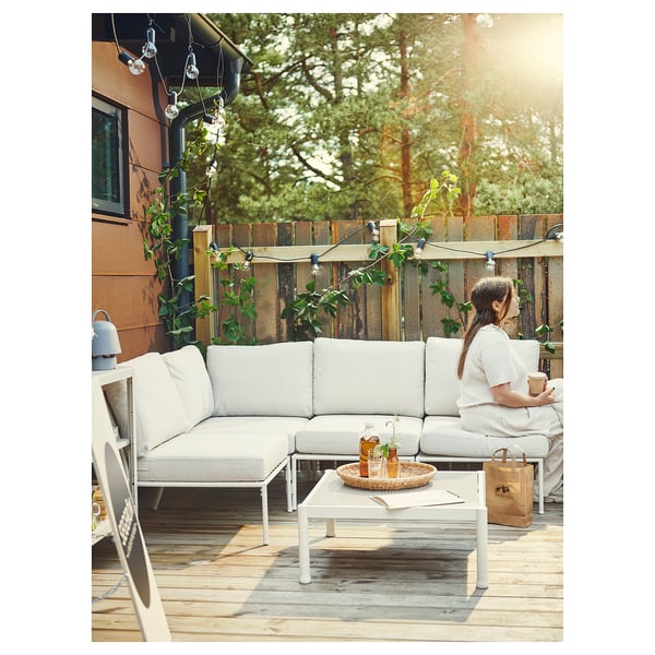Patio extérieur avec canapé SEGERÖN blanc. Femme assise, dégustant un café. Guirlandes lumineuses d’ambiance et terrasse en bois. Sac brun sur la table.