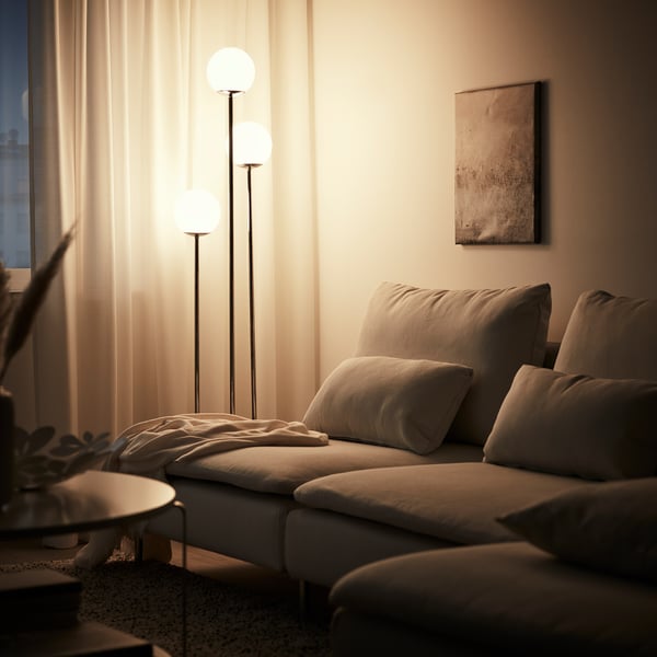 Salon confortable avec canapé beige, lampadaire éclairé, rideaux et plante.