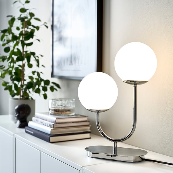 Lampe moderne SIMRISHAMN avec finition en verre et chrome. Il a des abat-jour sphériques, une base incurvée et se trouve sur une surface blanche à côté de livres et d’une plante.