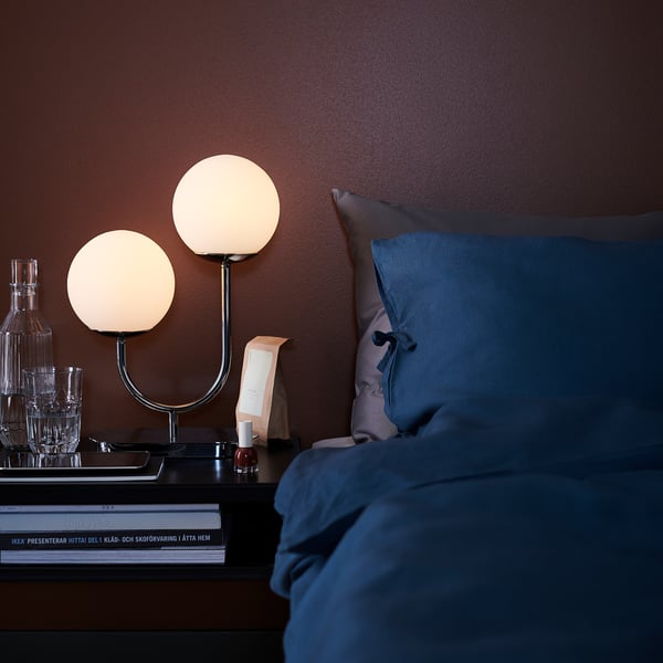 Chevet avec oreiller bleu, lampes à double globe, verre, chrome, livres et bouteille.