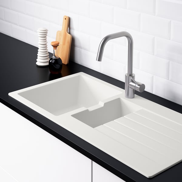 Évier de cuisine moderne avec égouttoir réversible, débit d’eau en pente, deux trous de robinetterie, comptoir blanc, noir, robinet argenté, planche à découper en bois.