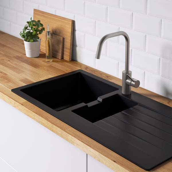 Évier de cuisine noir avec égouttoir, robinet en acier inoxydable, comptoir en bois, dosseret en brique blanche, bouteille, plante, planche à découper.