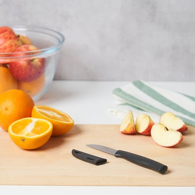 Une planche à découper en bois avec un couteau d’office, des moitiés d’orange, des tranches de pomme et un bol de pommes. Le couteau a un manche noir et un couvre-lame.