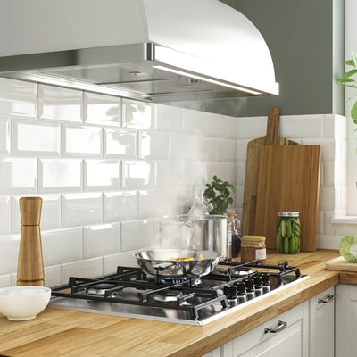 Cuisine moderne avec cuisinière à gaz, conduit en acier inoxydable, comptoir en bois, carreaux blancs, bol, bocal avec des cornichons, planche à découper et plante en pot.