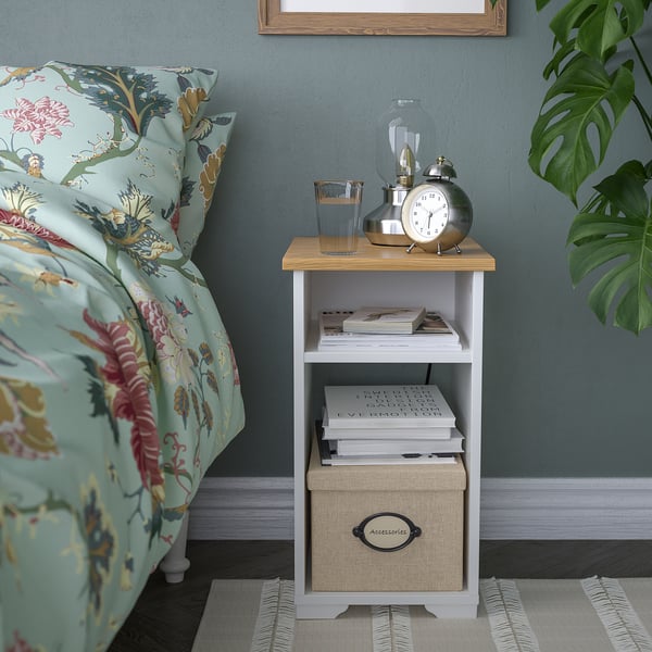 Une table d’appoint SKRUVBY avec un plateau en bois blanc et blanc, contenant des livres, une horloge et une lampe. À côté d’un lit avec une litière fleurie.