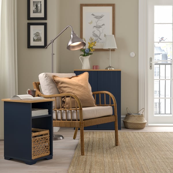 Coin confortable avec table d’appoint bleu marine SKRUVBY, chaise en osier, lampe et œuvre d’art. La couleur foncée des tables contraste avec un décor clair, s’adaptant parfaitement à côté de la chaise.