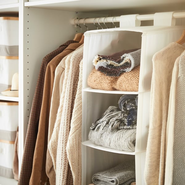 Placard bien organisé avec des vêtements beiges ; une unité de rangement suspendue étiquetée SKUBB en blanc contient des pulls pliés.