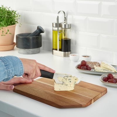 Personne tranchant du fromage sur une planche à découper en bois sur un comptoir de cuisine. À proximité se trouvent des raisins, des craquelins et des condiments.