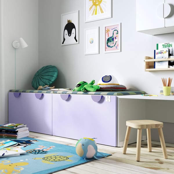 Chambre d’enfant avec coffre à jouets SMÅSTAD, violet clair, rectangulaire, range soigneusement les jouets. Il est plus grand que le tabouret à côté, se glisse sous l’art mural suspendu.