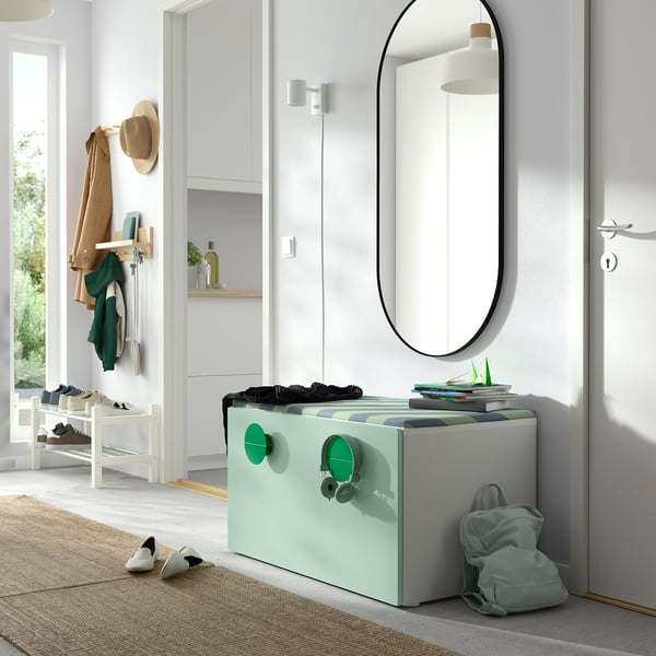 Entrée avec coffre à jouets SMÅSTAD vert menthe, livres et sac à dos ; murs blancs, miroir rond, patères et étagère à chaussures.