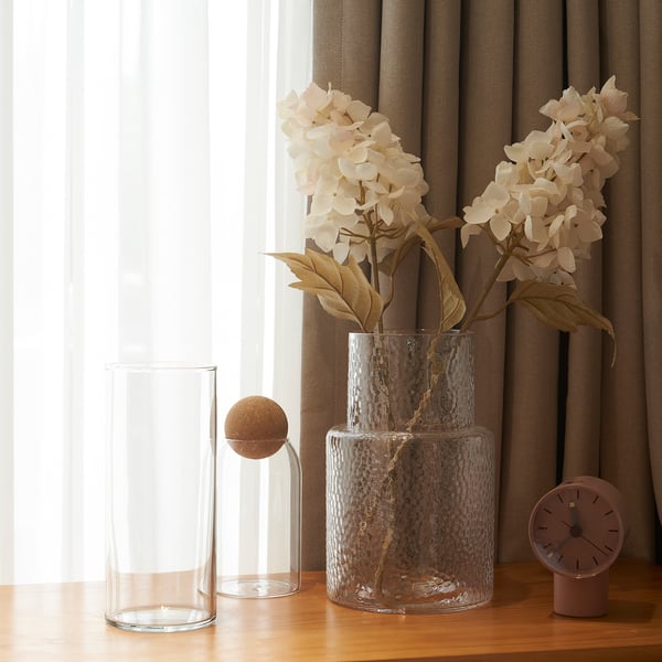 Des vases transparents et des fleurs artificielles aux pétales beiges et aux tiges brunes sur une table en bois à côté d’une fenêtre avec des rideaux.
