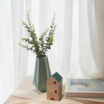 Décor minimaliste : vase vert à l’eucalyptus, maison en bois, rideaux transparents.