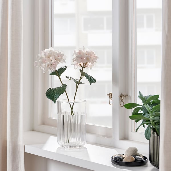 Décor de rebord de fenêtre : deux vases, l’un avec des fleurs rose clair, l’autre avec des feuilles vertes. Un bol de pierres lisses se trouve entre eux.