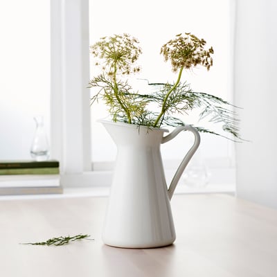 Vase en forme de cruche en céramique blanche contenant des fleurs séchées, posé sur une surface de couleur claire à côté d’une fenêtre, avec des tiges supplémentaires sur le sol.