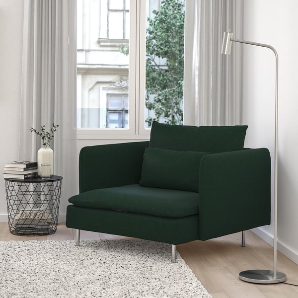Chaise SÖDERHAMN verte confortable près de la fenêtre. Sièges modernes et profonds, pieds en métal. Table d’appoint bien rangée avec des livres et un vase. Tapis moelleux en dessous. Lampadaire à droite.