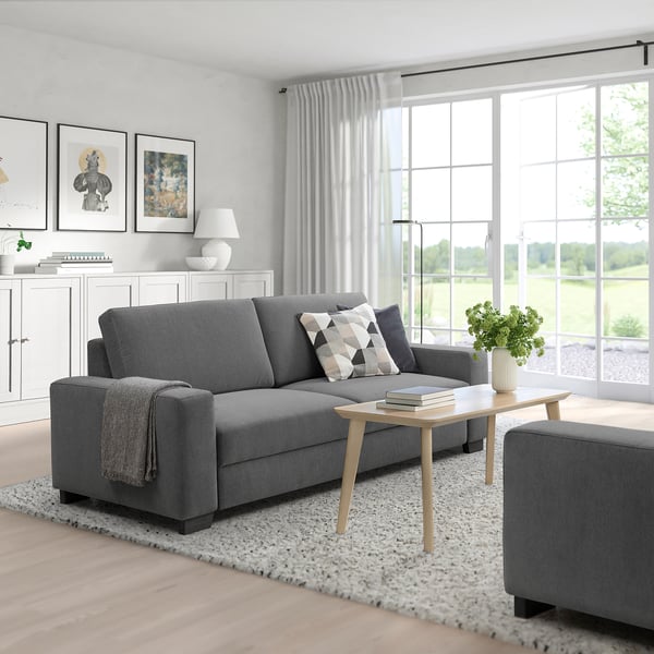 Salon moderne avec canapé SÖRVALLEN gris, pouf assorti, table basse en bois, rideaux blancs et grandes fenêtres offrant une vue verdoyante.