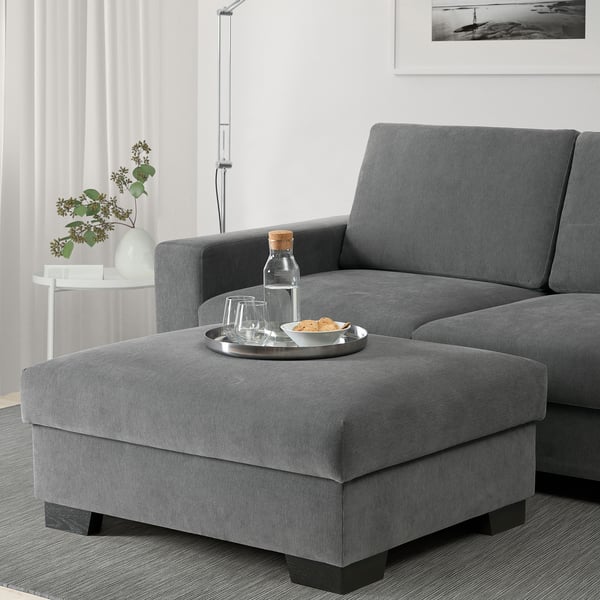 Salon moderne avec canapé en L gris, repose-pieds utilisé comme siège avec des collations, murs blancs, lampadaire et fleurs.