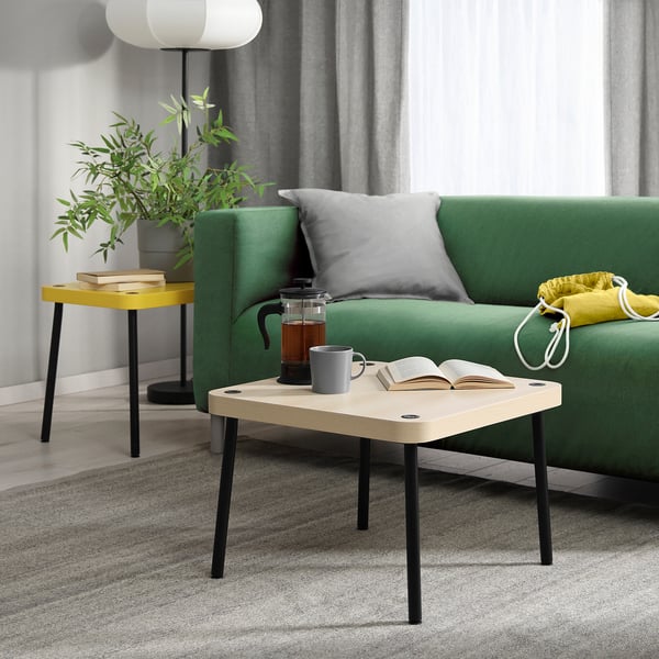 Petite table d’appoint SONHULT en blanc et noir, design minimaliste moderne, contenant une cafetière, une tasse et un livre à côté d’un canapé vert.