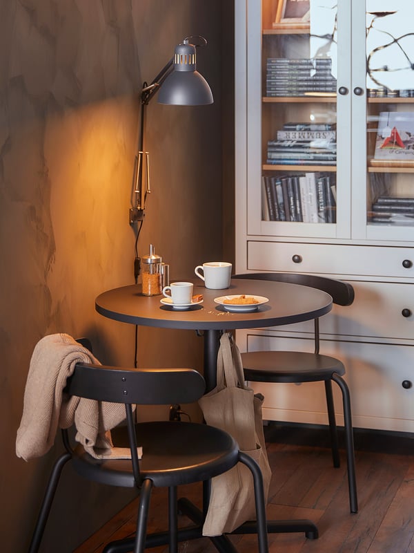 Coin confortable avec table ronde noire STENSELE, deux chaises, lampe, tasses et étagère.