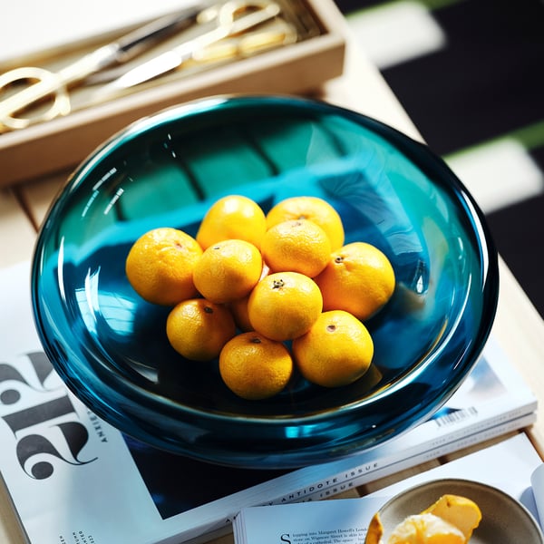 Un bol en verre bleu rempli de petits fruits ronds se trouve sur des magazines. La forme organique des bols en fait une pièce décorative.