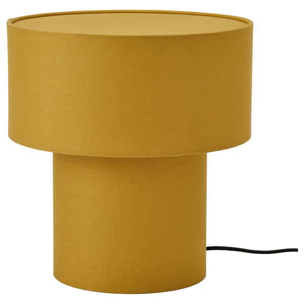 Une lampe de table STOCKHOLM 2025 jaune moutarde avec une forme cylindrique et un abat-jour en tissu de lin, donnant une lumière douce et chaude.