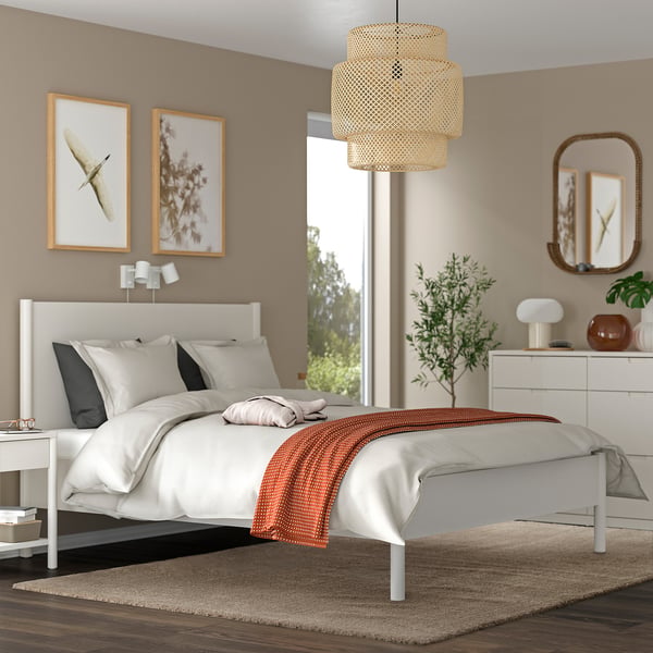 Chambre moderne avec murs beiges, lit blanc, art, lampe, plante, miroir et tapis brun chaud.