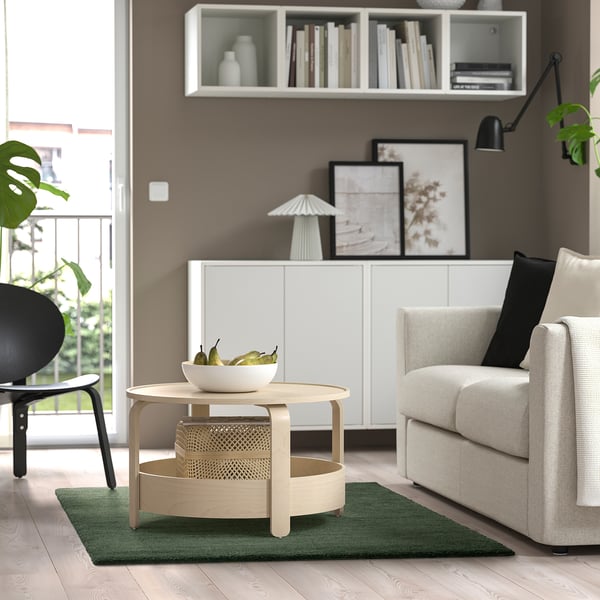 Salon moderne avec canapé gris, table en bois, tapis vert, chaise noire, armoire blanche, étagère, lampe noire, grande fenêtre.